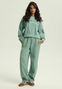 Miętowe dresy i pasująca krótka bluza z ribbowanymi mankietami, z nadrukowanym logo i kieszeniami bocznymi. Brązowe sneakersy dopełniają ten zestaw.