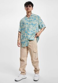 Kortermet, lysegrøn skjorte med blå palmetræmønster; beige cargo-bukser med sidelommer; hvide sneakers.