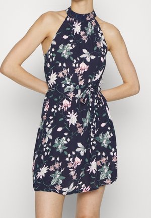 Kvinde iført ærmeløs marineblå blomstret kjole med halterneck og bundet talje, stående med hænderne på hofterne.