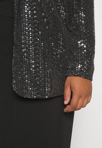 La mano destra appoggiata accanto a pantaloni neri e a una maglietta nera a maniche lunghe decorata con paillettes argentate.