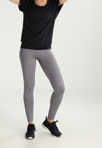 Černý krátký rukáv, šedé legíny s jemným zvířecím vzorem a černé sportovní boty. Legíny mají bílý logo akcent.