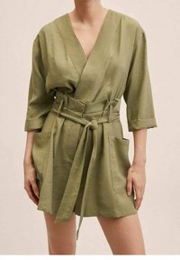 Vestido cruzado de lino en verde oliva con escote en V, mangas tres cuartos, cinturón y bolsillos delanteros. Textura suave con detalles de costuras mínimas.