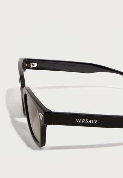 Μαύρα ορθογώνια γυαλιά ηλίου με σκοτεινούς φακούς, λογότυπο Versace στο πλάι και μικρό έμβλημα Medusa κοντά στη μεσόφυση, σε λευκό φόντο.