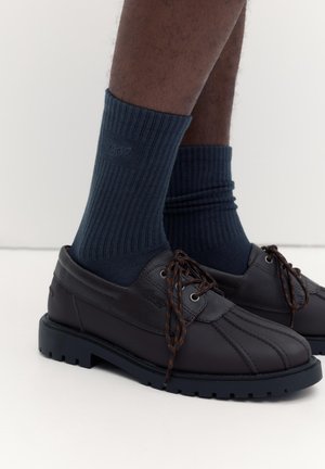 Sötétbarna bőrből készült, fűzős cipők, amelyeket bordázott sötétkék zoknival viselnek, egy személy alsó lábszárán, egyszerű világos háttér előtt.