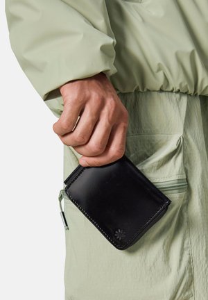 Portefeuille en cuir noir tenu à la main, présentant une texture lisse, des coins arrondis et un détail de surpiqûre décorative. Fond décontracté avec tenue vert clair.