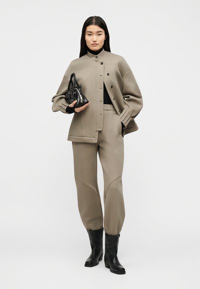 Ženska, oblečena v taupe velikansko jakno in hlače, črn puli in škornje, drži majhno črno torbico na ozadju z enobarvno podlago.