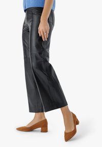 Ženska v črnih usnjenih kratkih hlačah in rjavih semiš čevljih s blok peto, stoji na beli podlagi, pri čemer je vidna ena roka.