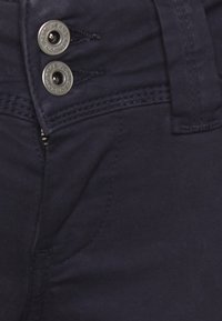 Tmavě modré džíny z denimu s hladkou texturou, dvěma kovovými knoflíky v pase a detailním šitím podél pásku.