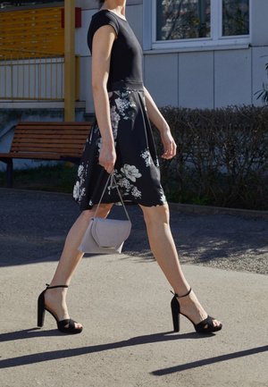 Zwarte jurk met bloemenprint, zwarte enkellaarsjes met riempje en een kleine lichtgrijze handtas. Op de achtergrond een bankje en een gebouw.