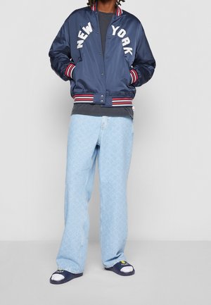 Persona che indossa una giacca varsity New York blu navy, t-shirt nera, jeans azzurri con motivo, calzini bianchi e sandali a ciabatta blu navy.