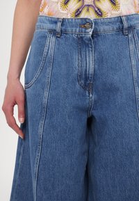 Pantalon en denim taille large en denim bleu clair. Comprend des poches avant, des coutures visibles, une fermeture à bouton et un design taille haute.