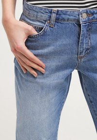 Světle modré džíny z džínsoviny s mírně vybledlou texturou, tradičním pětiokrovým designem a hardwarem v měděném odstínu.