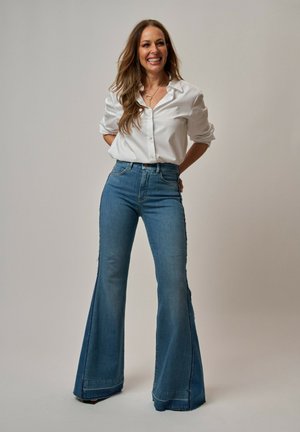 Mujer sonriente con cabello largo que lleva una camisa blanca y vaqueros azules acampanados de cintura alta, de pie con las manos detrás de la espalda.