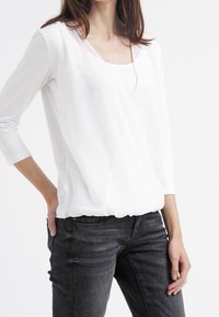 Haut blanc à manches longues avec un col en V, fabriqué en tissu lisse avec une base froncée, associé à un jean fitted gris foncé.