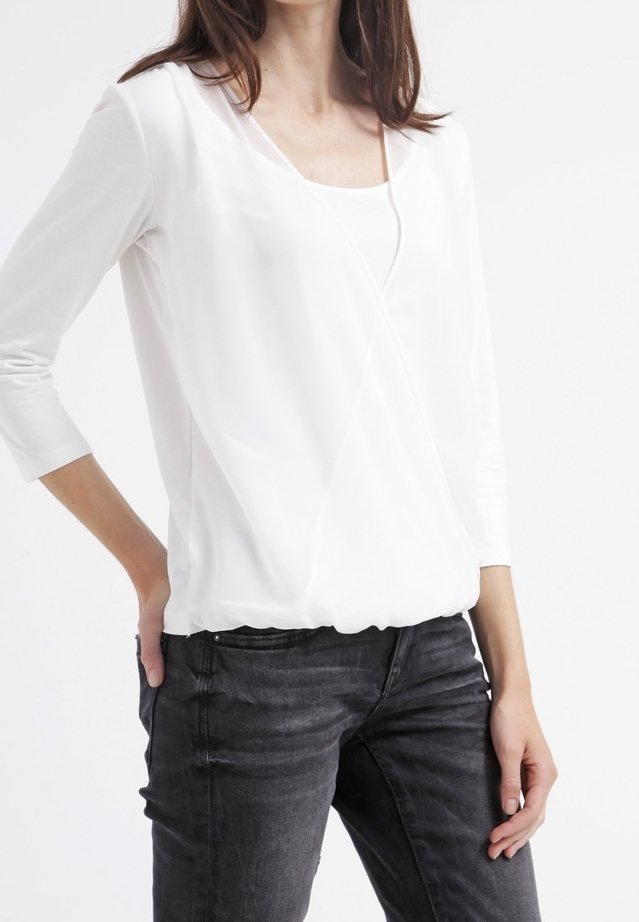 Haut blanc à manches longues avec un col en V, fabriqué en tissu lisse avec une base froncée, associé à un jean fitted gris foncé.