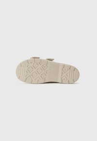 Beige sandaler med geometrisk såle, gummimateriale, to justerbare stropper og flat profil som er egnet for hverdagsbruk.