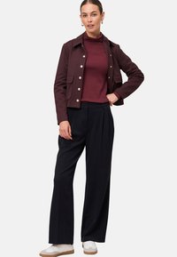 Woman wearing a maroon buttoned jacket over a maroon turtleneck, black wide-leg pants, and white sneakers, standing with one hand on hip.