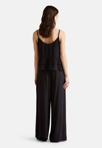 Zwarte, mouwloze jumpsuit met een uitlopende onderkant. Bevat gelaagde ruches rond de taille en verstelbare spaghettibandjes. Zachte textuur.
