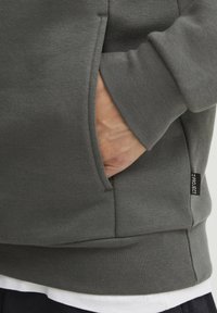 Gray sweatshirt with a textured fabric, featuring a side pocket and a small tag on the hem. Cuffs softly fitted at the wrists.