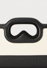Handtasche in Creme und Schwarz mit einem markanten Griffdesign und strukturierten Akzenten, sowie einem prägedruckten Logo an der Basis.