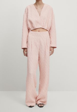 Femme portant un crop top texturé rose pâle à manches longues et un pantalon large taille haute avec des chaussures noires à bout ouvert sur fond blanc.