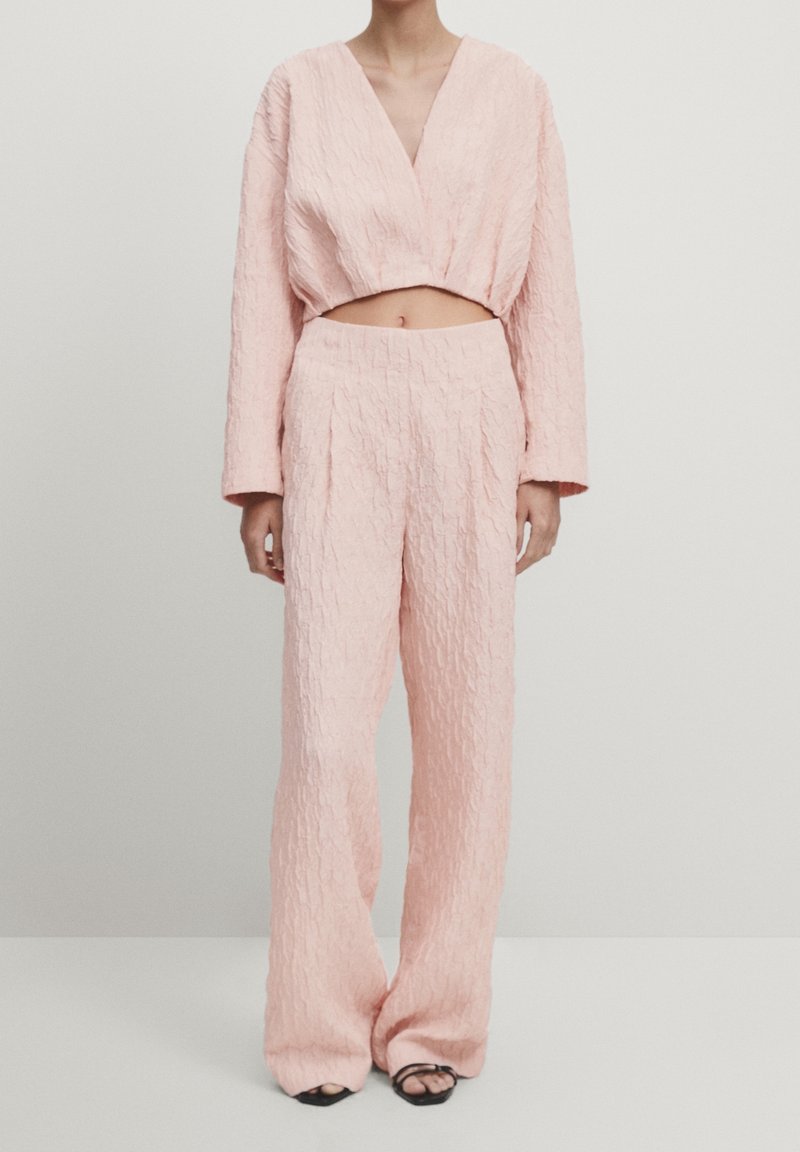 Femme portant un crop top texturé rose pâle à manches longues et un pantalon large taille haute avec des chaussures noires à bout ouvert sur fond blanc.