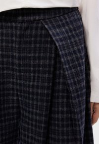 Mornarice karirane hlače od vune, s teksturiranim materijalom, širokim nabranim dijelovima i prilagođenim pojasom. Bočne džepove također su vidljive.