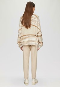 Chunky gebreide trui met beige en bruine strepen, ruime pasvorm en dropped schouders, gecombineerd met lichtbeige broek en crèmekleurig schoeisel.