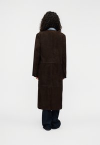 Καφέ μακρύ παλτό από σουέντ με ευθεία γραμμή, ορατές ραφές και πίσω άνοιγμα. Φορεμένο πάνω από μπλε τζιν παντελόνι. Υφή με απαλό φινίρισμα.