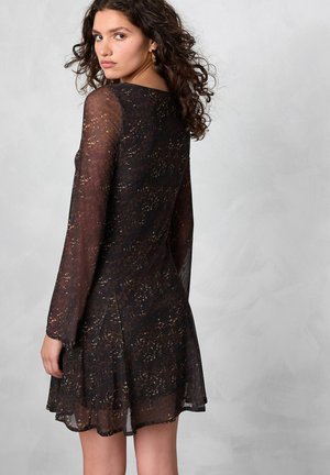 Robe noire transparente à manches longues, présentant un motif texturé de points dorés et marron. Jupe évasée avec des fentes sur les côtés à l'ourlet.