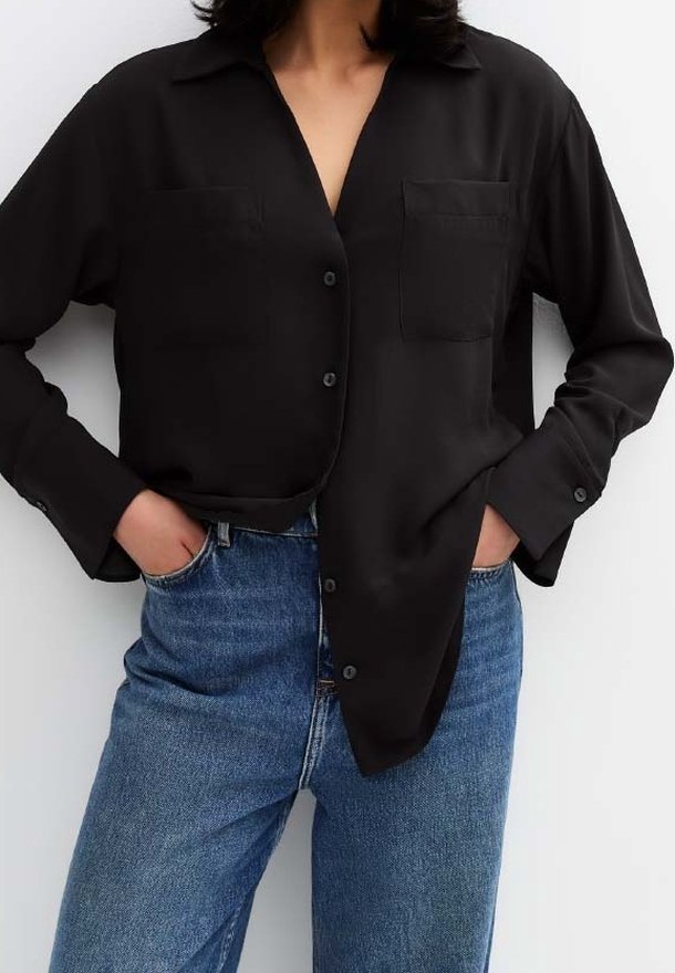 Femme portant une chemise noire ample boutonnée, rentrée de manière inégale dans un jean bleu taille haute, mains dans les poches.