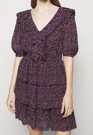 Femme portant une robe fleurie violette avec des manches bouffantes, un col en V profond, un bord à volants et des couches de jupe à niveaux sur un fond uni.