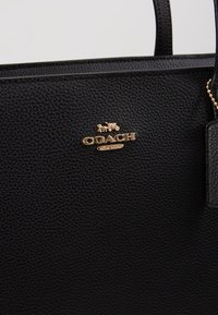 Musta nahkainen olkalaukku, jossa on teksturoitu pinta, kullansävyinen "COACH"-logo ja nimilappu. Hihnat ovat tukevat ja hyvin ommellut.