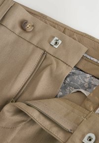 Pantaloni marroni chiari con pattina e chiusura a bottone. Interno con una fodera a motivo floreale; cuciture e texture ben rifinite.