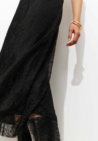 Robe en dentelle noire avec un tissu transparent et texturé, présentant un ourlet asymétrique. Une main portant plusieurs bracelets en or est partiellement visible.