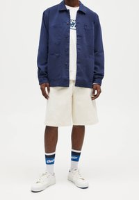 Marinblå knappup jacka, vit grafisk sweatshirt under, krämfärgade shorts och vita sneakers med blå detaljerade strumpor.