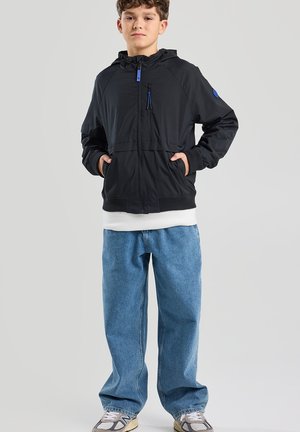 Zwarte lichtgewicht jas met capuchon, rits aan de voorkant en blauwe accenten, gecombineerd met losse blauwe spijkerbroeken en grijze sneakers.