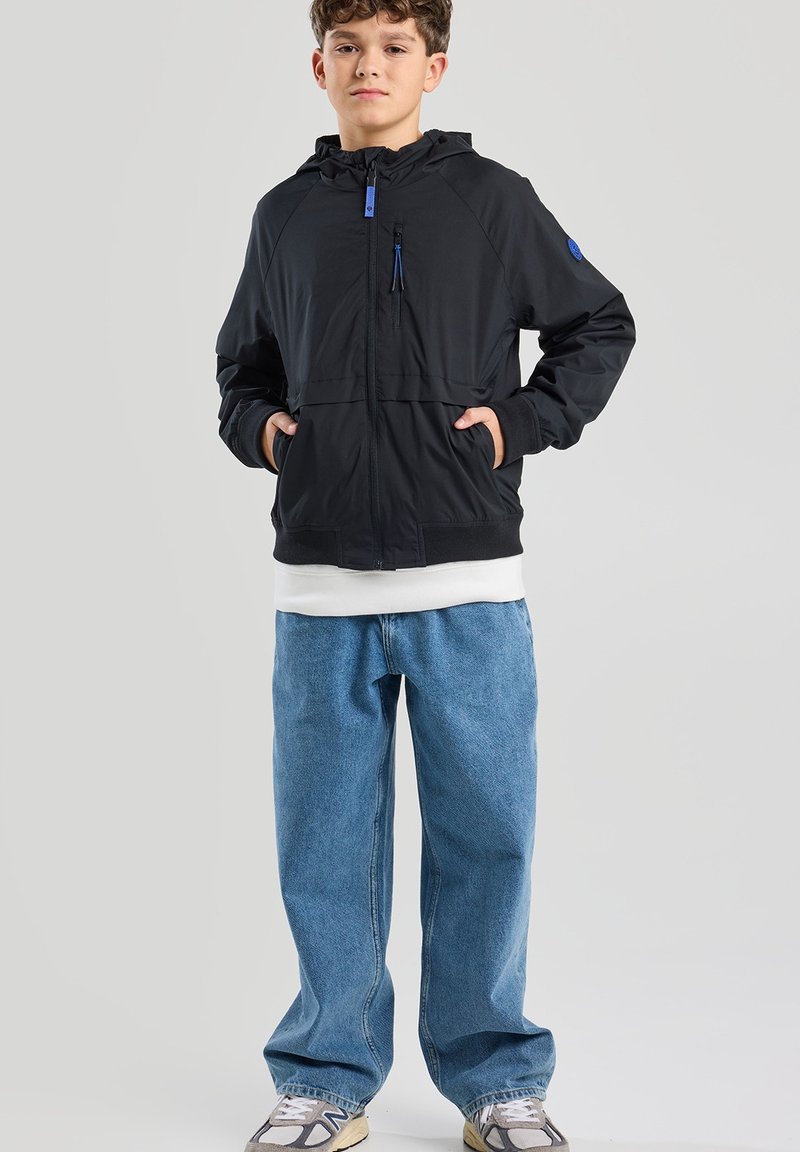 Musta kevyt takki hupparilla, vetoketjukiinnitys edessä ja sinisiä yksityiskohtia, yhdistettynä väljään sinimekkopariin ja harmaisiin lenkkareihin.