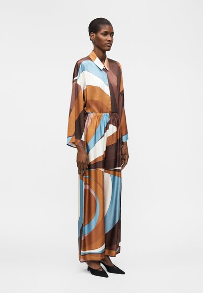 Γυναίκα που φοράει μακρύ, φαρδύ φόρεμα με καφέ, μπλε, λευκές και πορτοκαλί αφηρημένες μορφές, συνδυασμένο με μαύρα μυτερά παπούτσια.