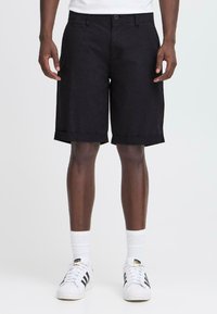 Person klädd i svarta knälånga shorts med uppvikta ben, vita strumpor och vita Adidas-sneakers med svarta ränder, som står mot en enkel bakgrund.