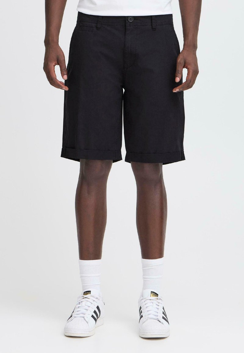 Person klädd i svarta knälånga shorts med uppvikta ben, vita strumpor och vita Adidas-sneakers med svarta ränder, som står mot en enkel bakgrund.
