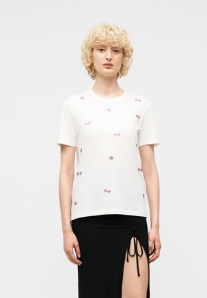 Γυναίκα με σγουρά ξανθά μαλλιά φοράει λευκό κοντομάνικο τοπ με μικρές μωβ βούλες και μαύρη φούστα με ψηλή σχισμή δεμένη με κορδόνια.