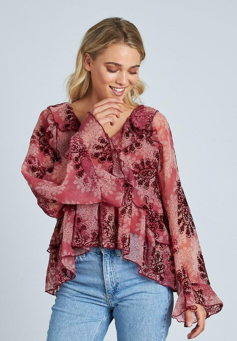 Blusa ligera y fluida en color malva con patrones florales oscuros, cuello con volantes y mangas de campana. Combinada con jeans de mezclilla claros.