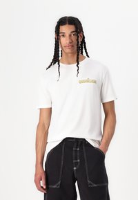 Hvit T-skjorte med korte ermer og Quiksilver-logo i gult på venstre bryst, brukt med svarte bukser med hvite sømdetaljer.