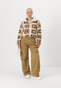 Kort, mönstrad jacka i pastellfärger med pälsfoder. Vida khaki cargobyxor med flera fickor, tillsammans med vita sneakers.