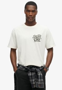 Camiseta corta de manga en color crema con el logo negro "SUPERDRY", combinada con pantalones negros y una camisa de cuadros gris atada a la cintura.