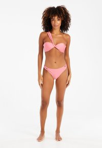 Růžový bikini set s topem na jedno rameno s uzlovým detailem a odpovídajícími spodními díly s bočními výřezky. Hladká textura, minimální kovové prvky.
