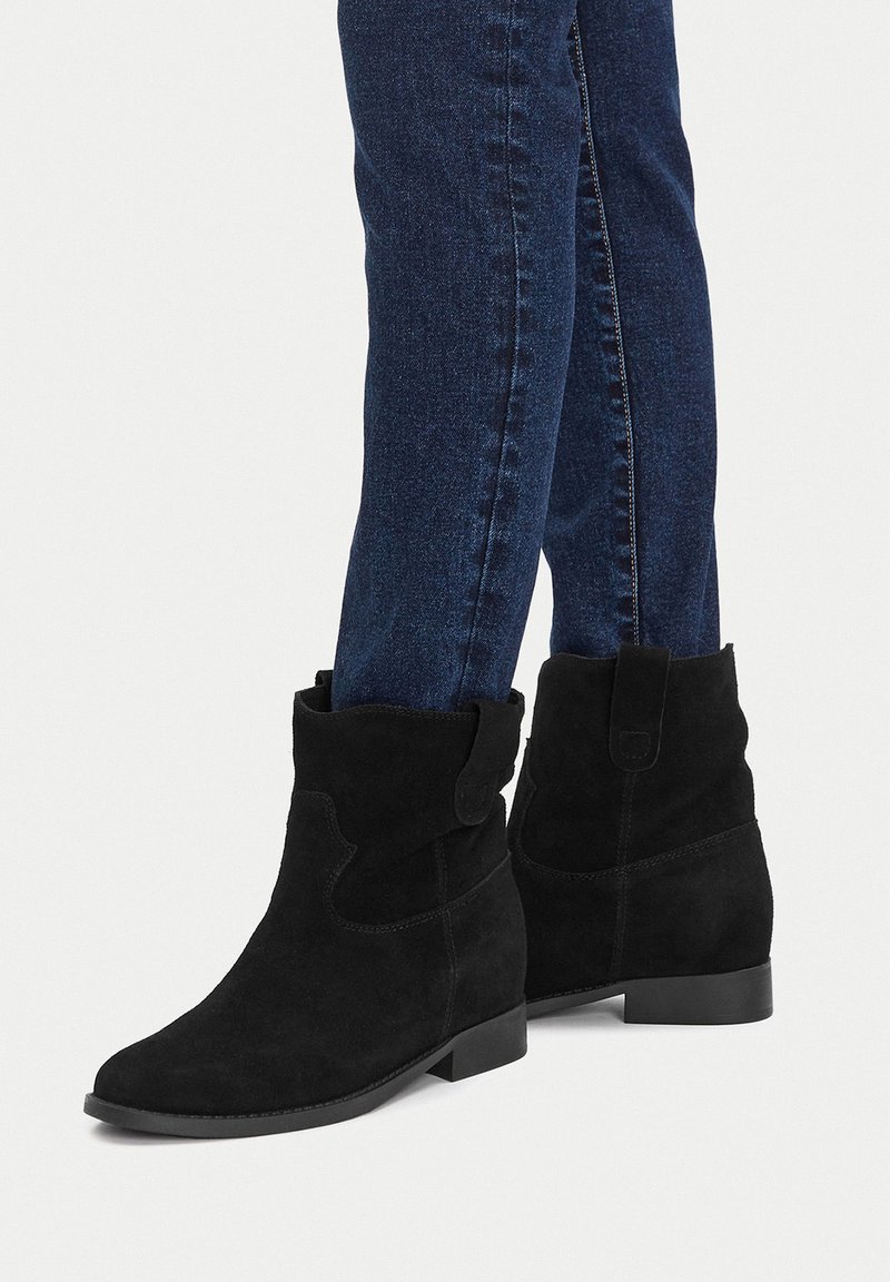 Botine negre din suede, cu toc mic, cu un manșon pliat și un detaliu de nasture. Asortate cu blugi skinny de culoare albastru închis.