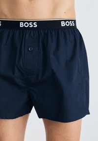 Sötétkék pamut boxer alsó, elasztikus derekkal, amelyen a "BOSS" márkaneve fehér színben szerepel. Egy gombos záródással rendelkezik.