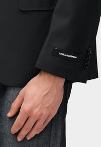 Μανίκι μαύρου σακακιού με ορατά κουμπιά και μια ετικέτα υφάσματος που αναγράφει "KARL LAGERFELD." Το υλικό είναι κομψό με απαλή υφή.